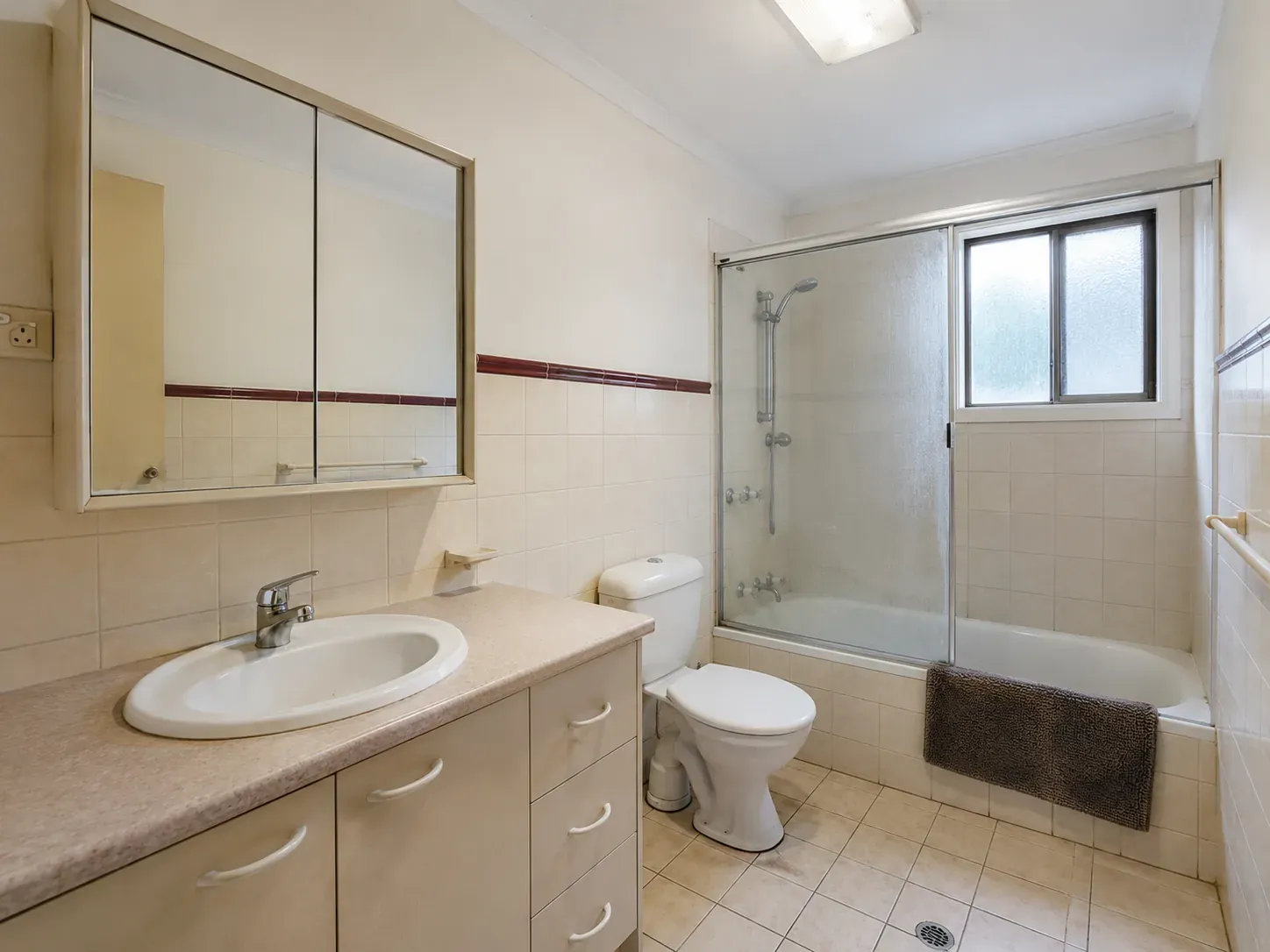 A tired 1990s Australian family bathroom with cream wall tiles, builder-grade laminate vanity, dated polished chrome tapware and shower-over-bath