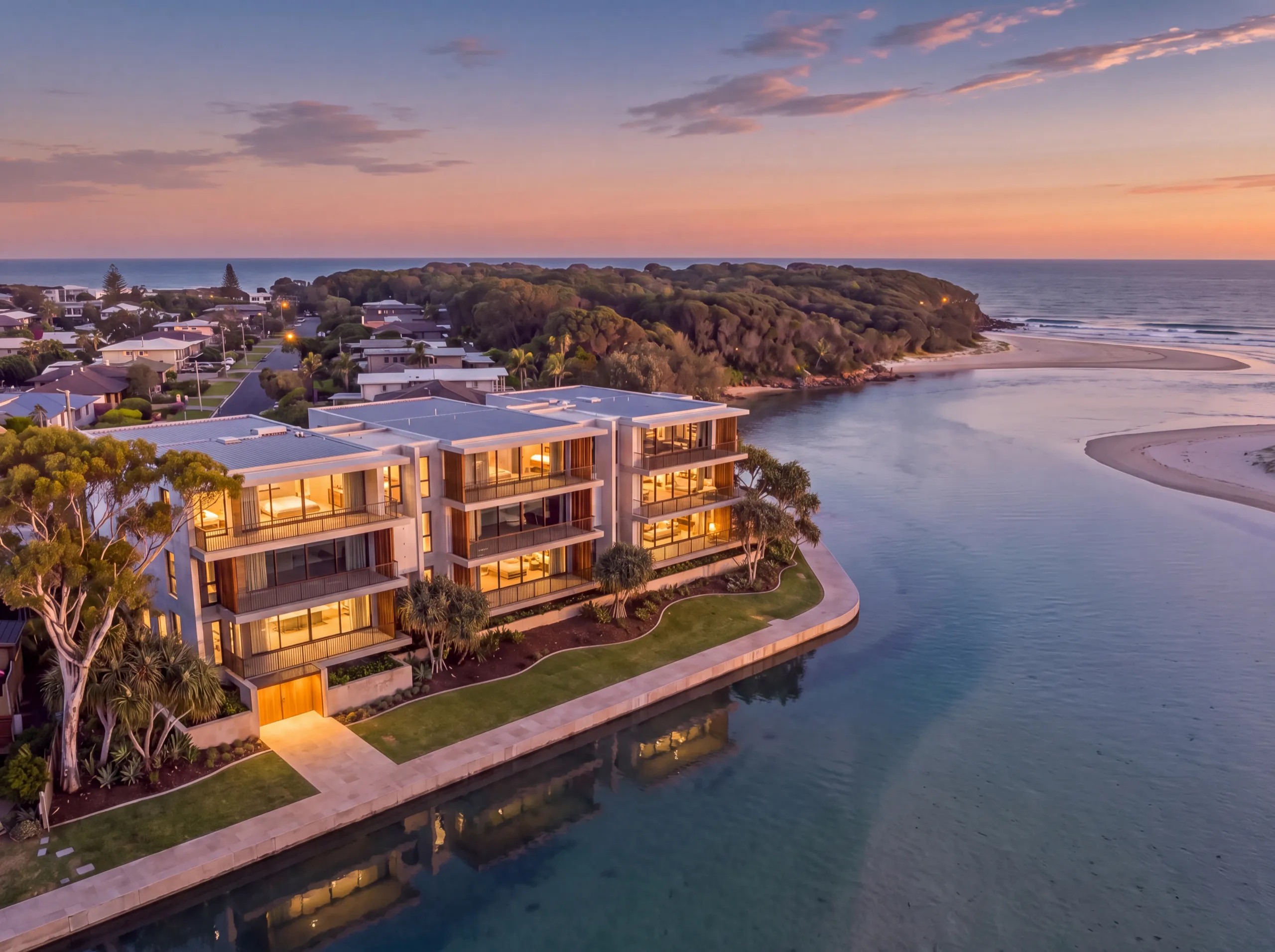 Aerial at dusk of a three-storey waterfront building on a tidal creek, the headland and the sea beyond